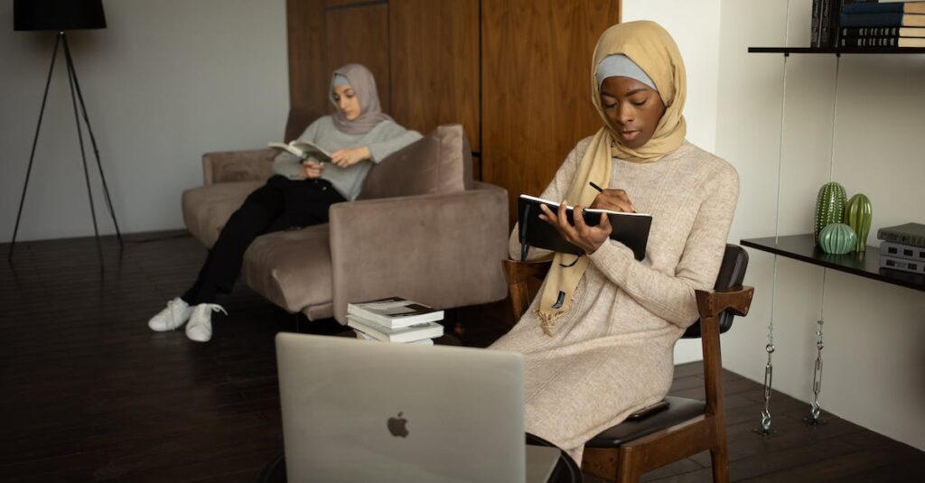 Focused young black Muslim female taking notes and watching tutorial via laptop near female friend reading book on sofa